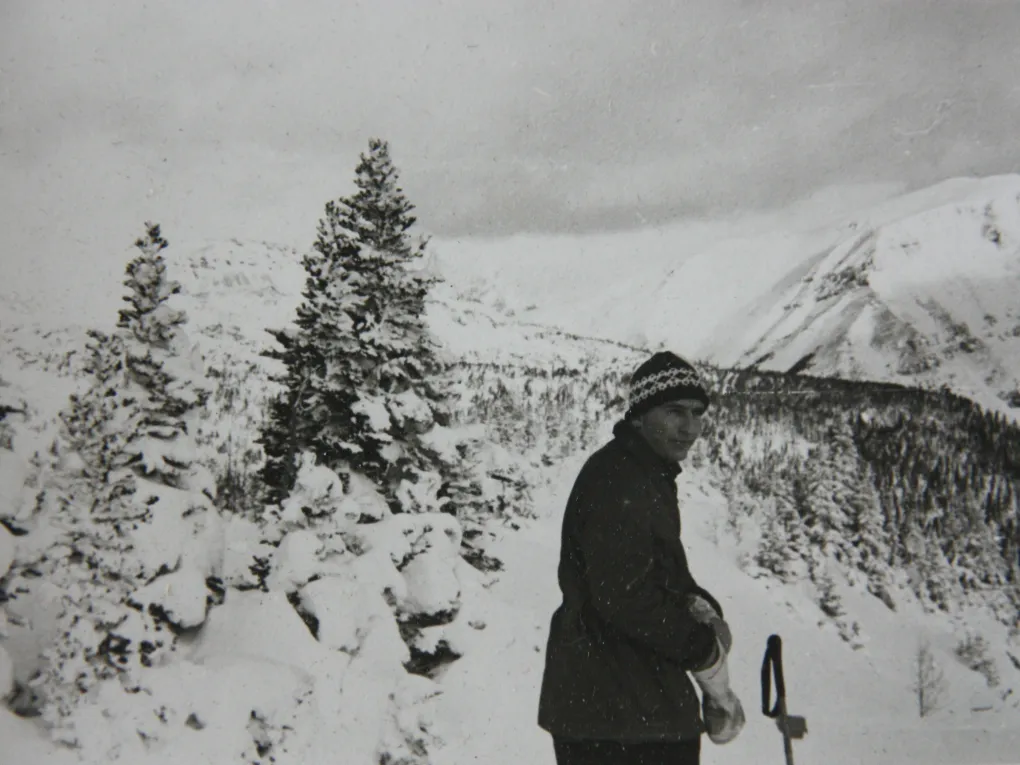 Gammelt svart-hvitt-bilde av en mann som står på ski. Snødekte fjell i bakgrunnen.