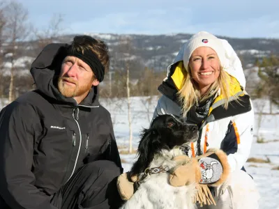 Dame ler mot kamera, mens hun koser med en hund. Mann ser seg til siden