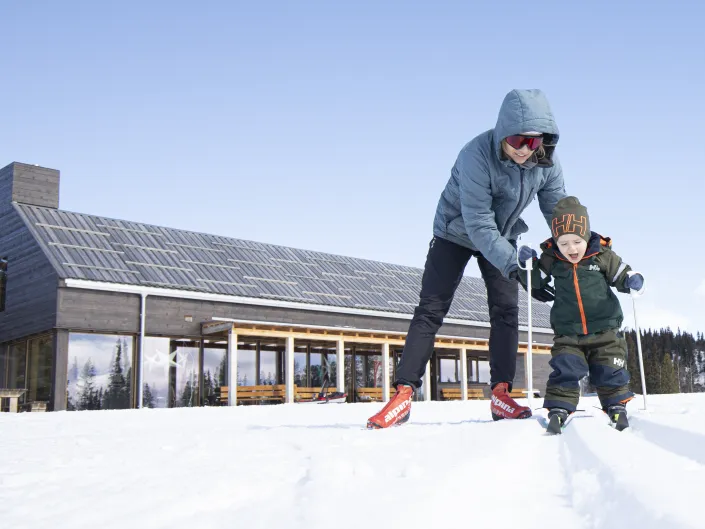 Et bilde av en mamma som hjelper barnet sitt med å gå langrenn, med gravsetstugu i bakgrunnen.