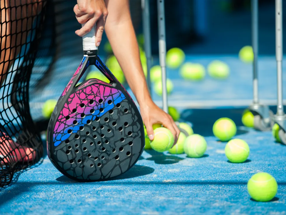 Bildet er av en hånd som plukker opp padel-baller med den ene håden, og holder rekkerten i den andre.