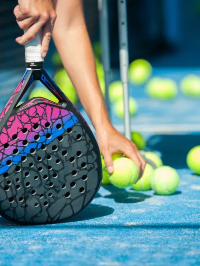 Bildet er av en hånd som plukker opp padel-baller med den ene håden, og holder rekkerten i den andre.