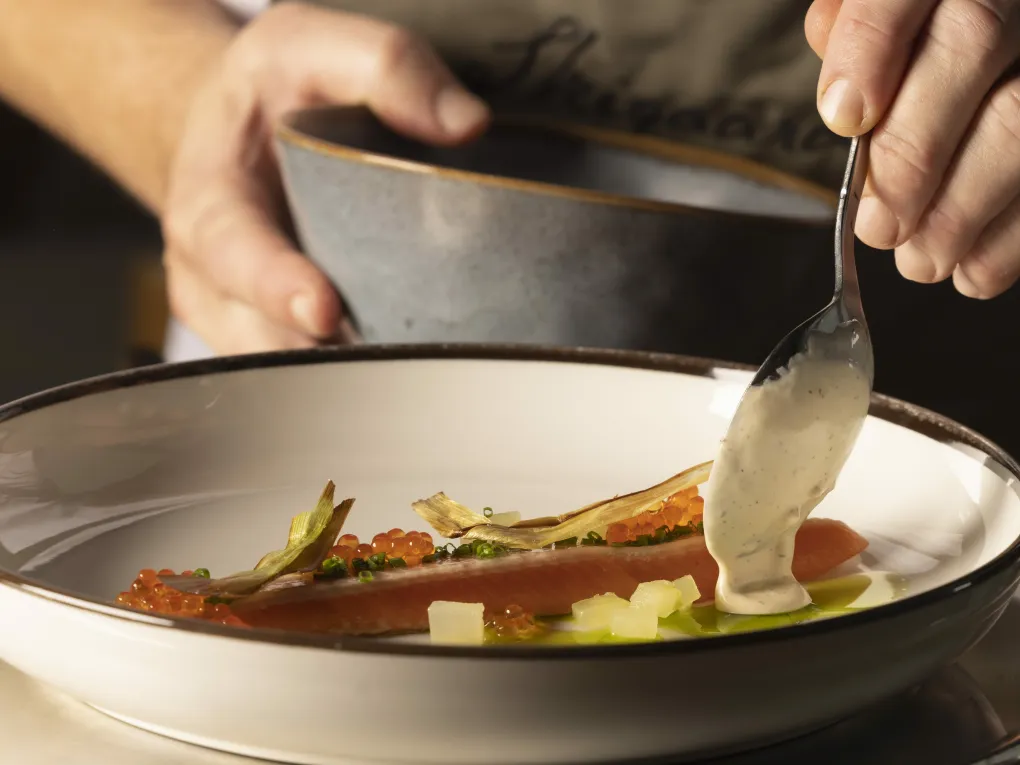 Fiskerett med delikat topping, og en hånd som bruker en skje til å helle saus.