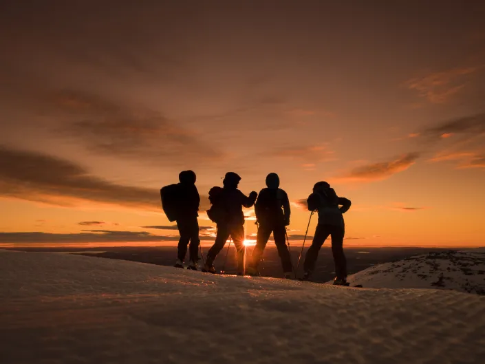 Personer med ski på beina i bakken, og solnedgangen i bakgrunnen.