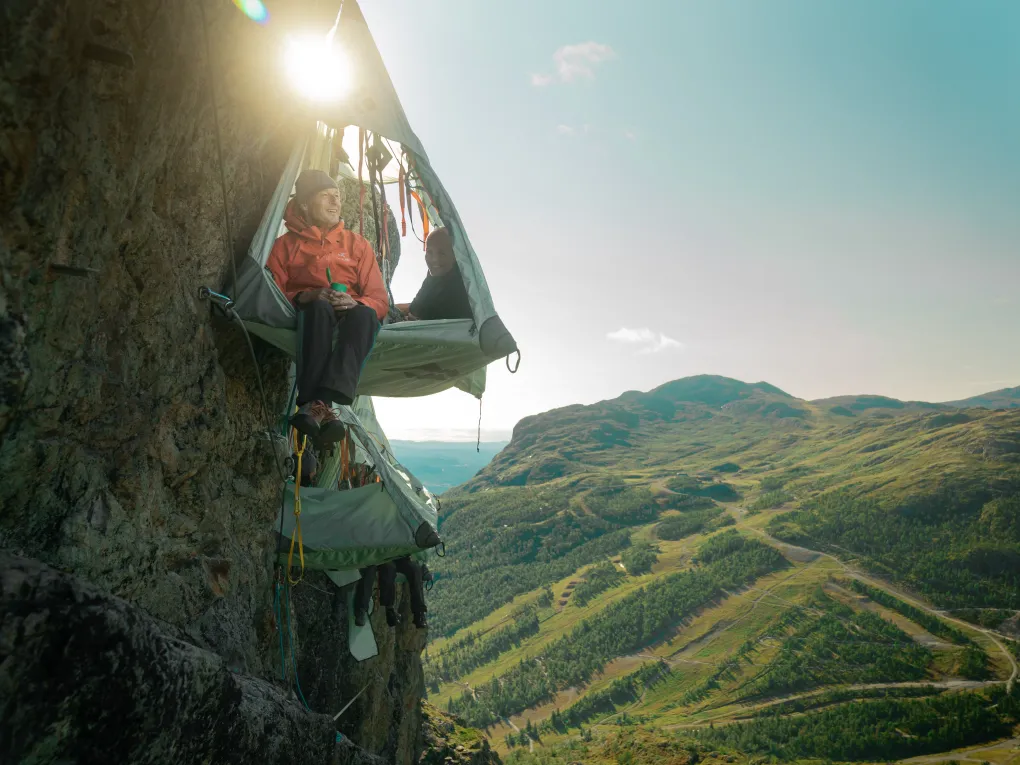 Mann som sitter med beina ut av skysleeping teltet, mens en dame ligger med hodet ut. I bakgrunnen av teltet er det flere andre telt og fjell.