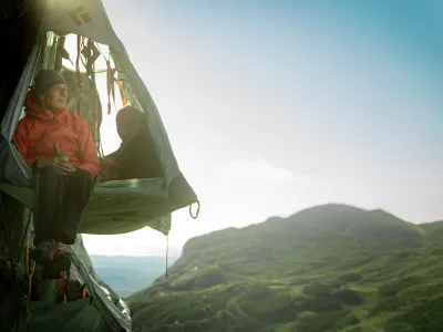 Mann som sitter med beina ut av skysleeping teltet, mens en dame ligger med hodet ut. I bakgrunnen er det fjell.