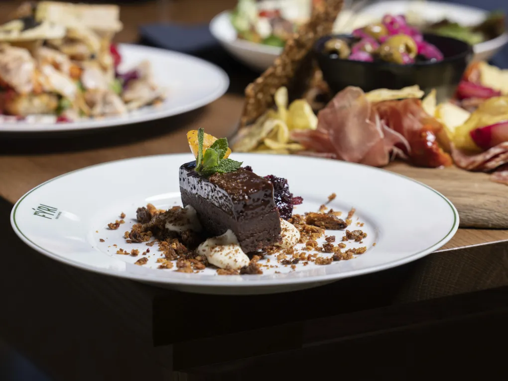 Her er det et bilde ev en sjokoladekake som er pyntet pent oppå en hvit tallerken, i bakgrunnen er det andre type desserter.