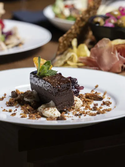 Her er det et bilde ev en sjokoladekake som er pyntet pent oppå en hvit tallerken, i bakgrunnen er det andre type desserter.