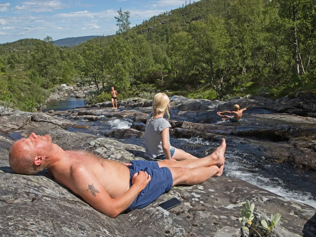 Mann soler seg, imens barna leker i elva.