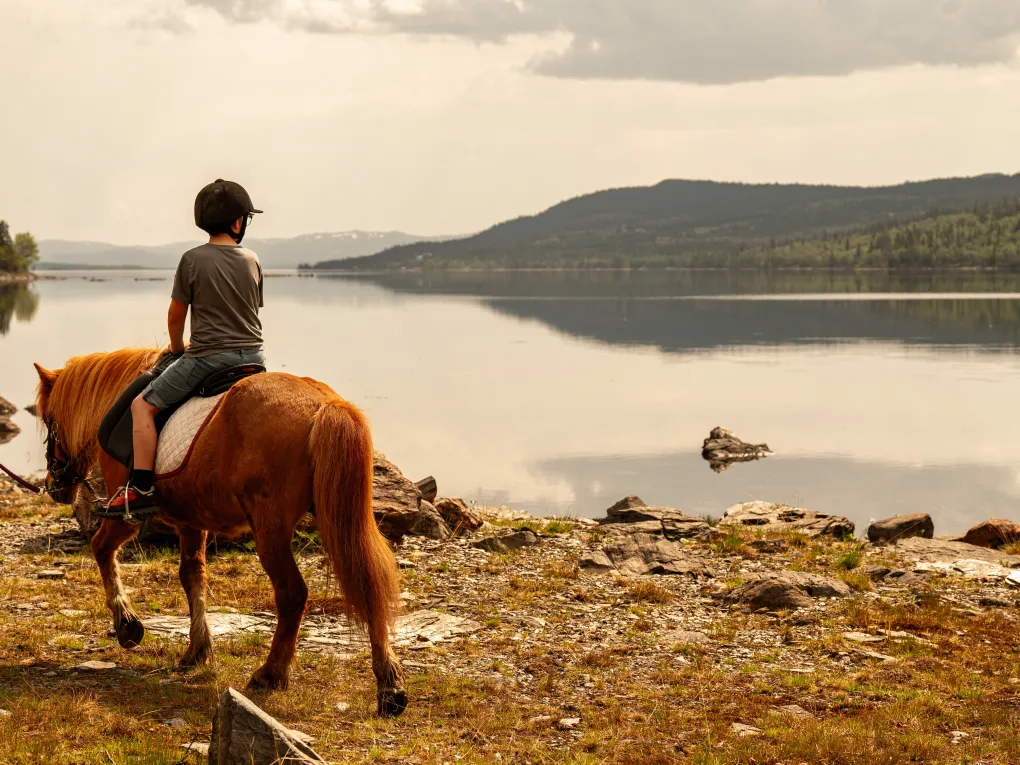 Gutt som rir på en brun hest, ved vannet, med fjell i bakgrunnen.