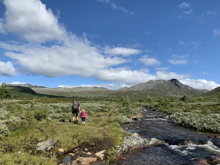 Mann og liten jente på tur i fjellet om sommeren, elv og fjell i bakgrunnen.