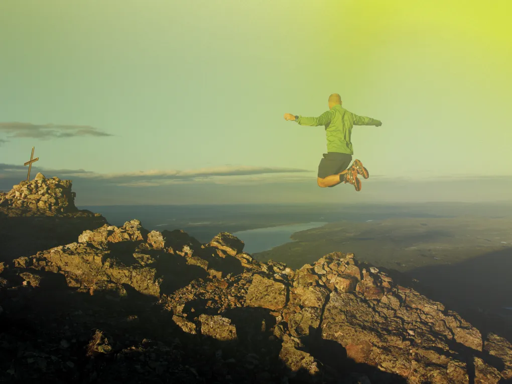 En mann som hopper på toppen av et fjell, han er ikledd grønnjakke.